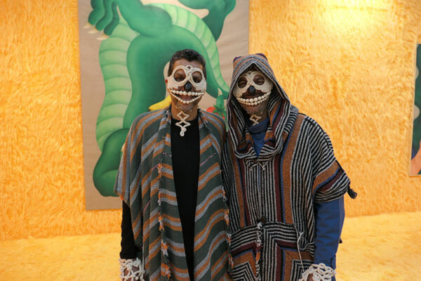 Two people in white skull masks pose for a phot in front of a large dragon painting.