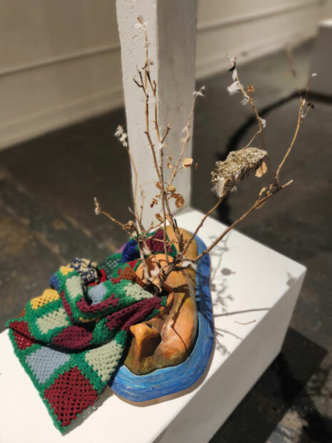 A clay sculpture with an afghan blanket and milkweed branches attached to it.