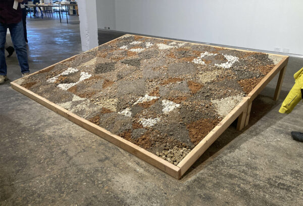 Sequences of different colored dirt are arranged in a quilt-like pattern on a tilted plinth lying on the floor.