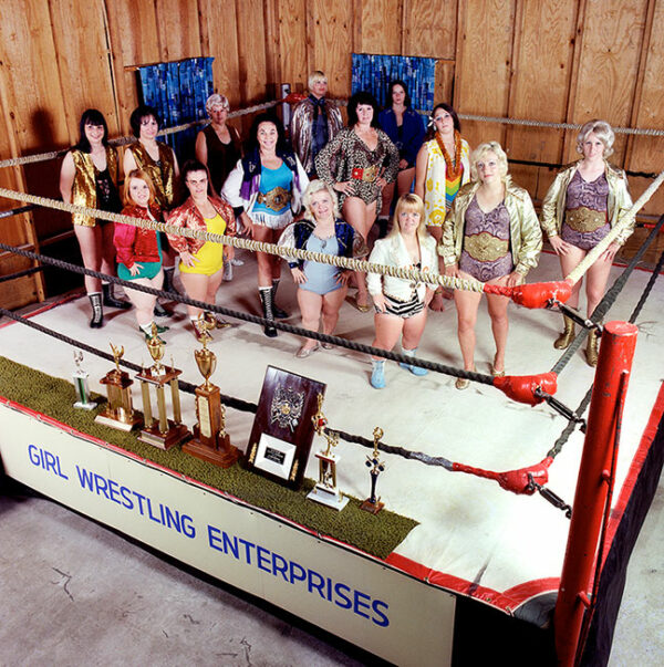 A large group of women stand inside a wrestling ring looking upwards into the camera.