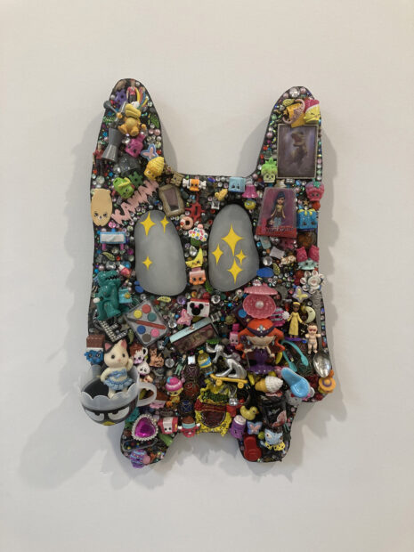 A cartoonish figure with upright ears and large eyes is hung on the wall. The sculpture consists of many miniature tchotchke's and plastic toys.