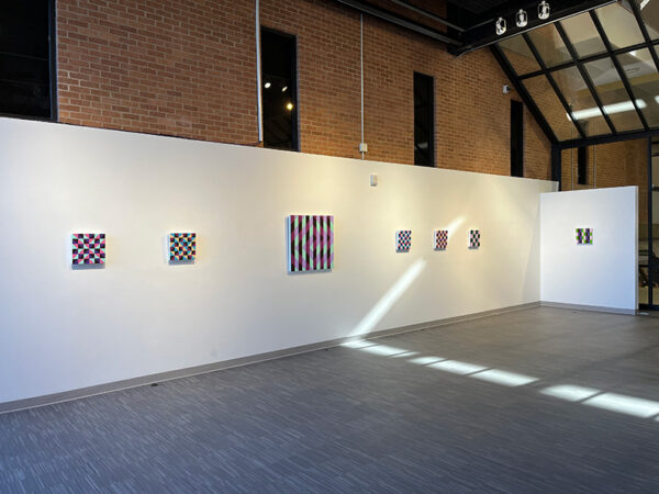 Seven hard-edged geometric abstract paintings hang on the wall of a high-ceilinged gallery.