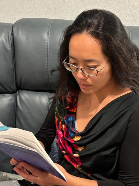 A woman with glasses in a black shirt on a grey couch reads a book.