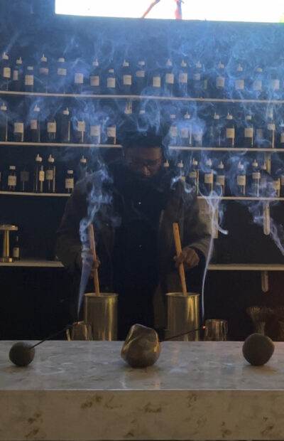 A man stands before a wall of labeled bottles holding two sticks inside brass bowls while several sticks of incense burn on the table before him.