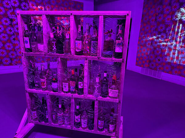 A wooden structure holing dozens of liquor bottles sits on the floor of a purple-lit gallery.