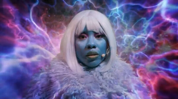 W oman with blue skin and white hair faces the camera with cosmic lights flashing behind her.