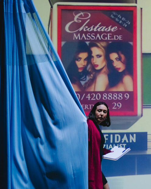 A woman in red holding a clipboard walks from behind a blue curtain in front of a large projection of an ad for erotic massage featuring three models.