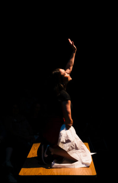 A woman kneels atop a desk while looking upwards with a fistful of paper and one outstretched hand towards the light.