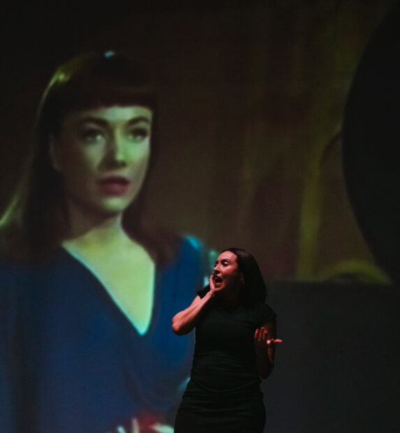 A performer stands before a screen with a giant projection of a woman in blue dress.