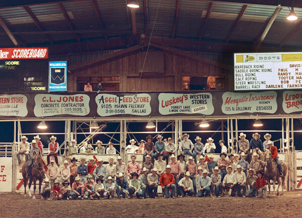 A large group of rodeo participants pose for a photograph inside an arena.