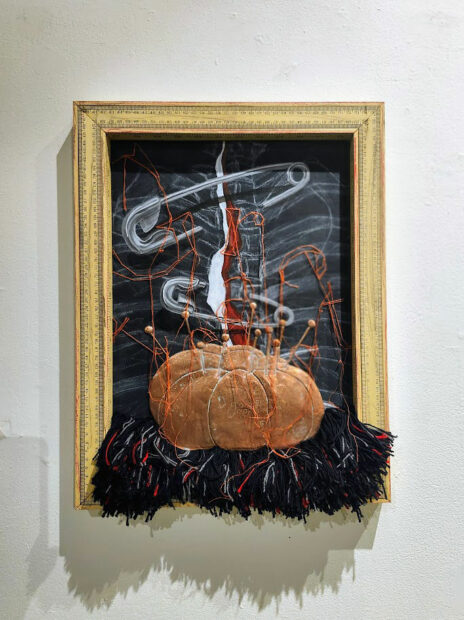 A framed multi-media work with a measuring tape frame and a low-relief pin cushion.