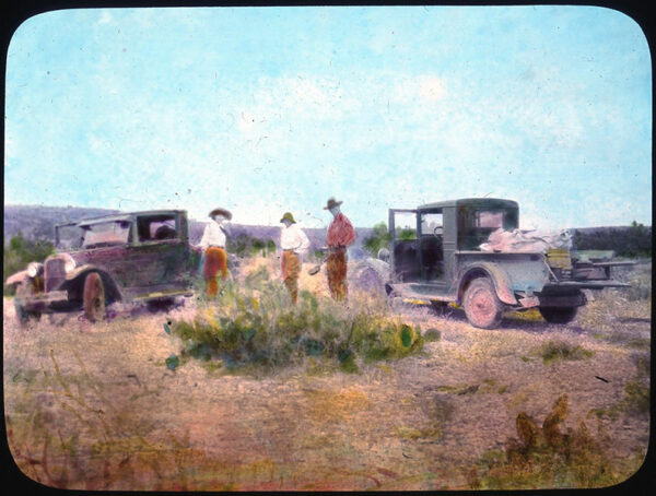 A hand-colored photograph depicts three people standing in the high desert plains next to two vehicles.