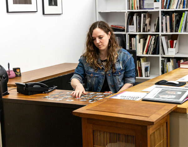 A woman with long hair wearing a jean jacket sits at a desk looking at photographs.