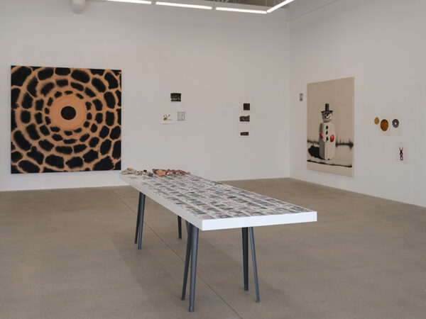 A gallery with a table covered in Polaroids sits before a wall with large paintings of a snowman and a black pattern.