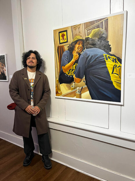 A man stands in front of a painting of a figure looking in a mirror.