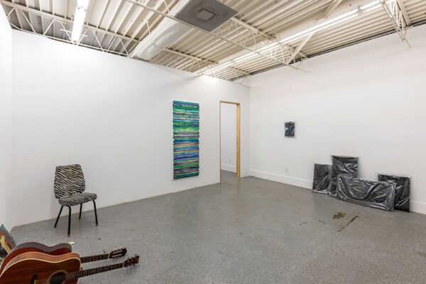 A gallery contains works on the wall and sculptural works leainng against the wall on the floor, as well as two guitars and a chair from a performance.