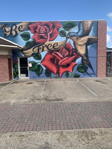 A photograph of the exterior of Be Free Gallery, featuring a large mural of roses.