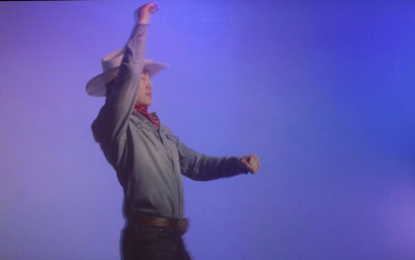 A cowboy gestures with his hands in front of a blue backdrop.