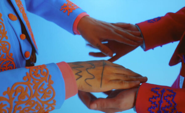 Two people in bright charro outfits with tattooed hands touch hands. 