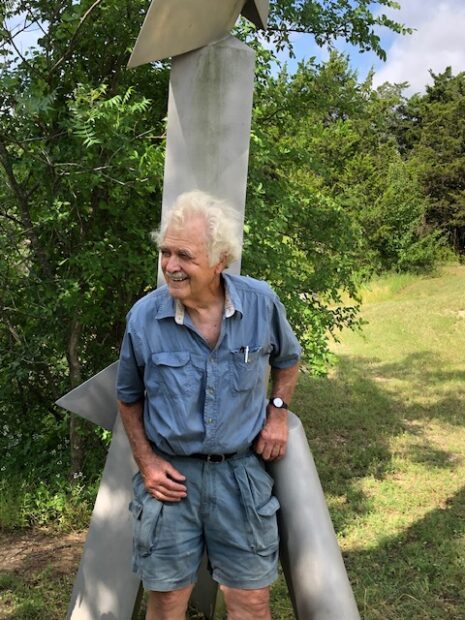 A photograph of artist Mac Whitney smiling as he stands near a large sculpture.