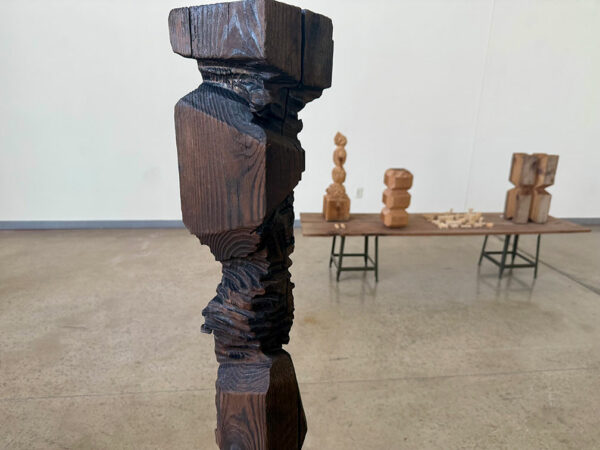 In the foreground a large, dark wooden sculpture stands before a table with smaller carved sculptures.