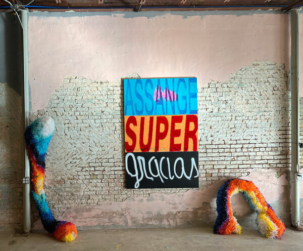 A painting with the phrase" Assange super gracias" on it hangs between two paper mache sculptures.