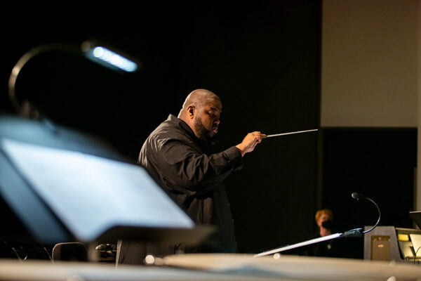 A man stands in a darkened room leading musicians with a conductor's baton in his hand.