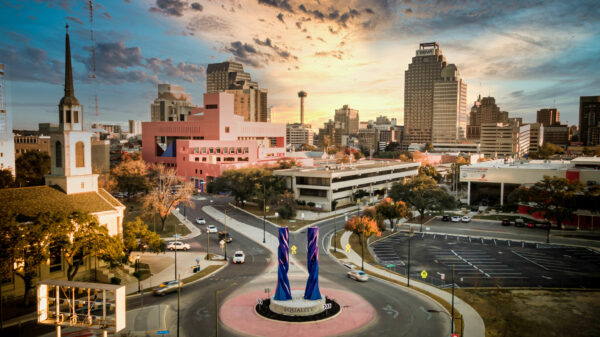 An aerial photograph of a roundabout in downtown San Antonio, featuring a skyline of buildings.