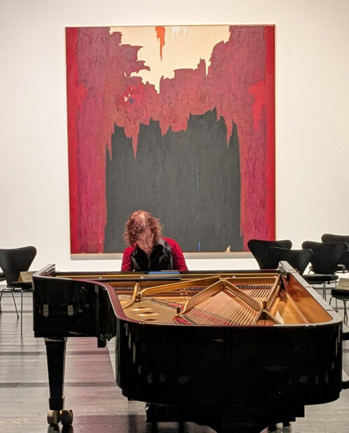 A woman sits at a grand piano in a gallery in front of a large abstract painting.