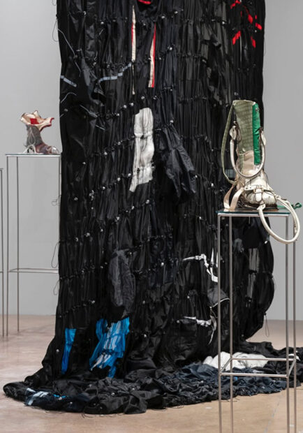 Several handmade boots stand atop pedestals before a large fabric piece in a gallery.