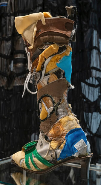 A boot, made from scraps of other garments sits atop a pedestal in a gallery.