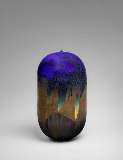 A bean-shaped ceramic sculpture glazed in some brilliant, glowing colors, and some neutral, flat colors.