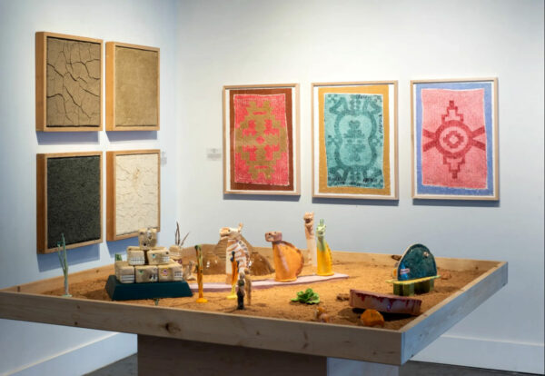 A tabletop tableau with small figures in a gallery with framed works on a wall.