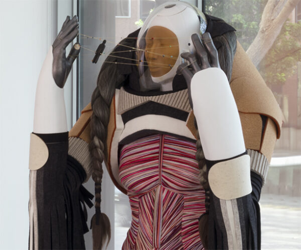 A photograph of a sculpture of a human dressed in a futuristic suit and holding a string in a cat's cradle formation.