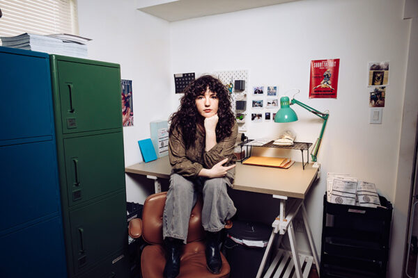 A woman sits on a desk in a studio with photographs pinned to the wall.