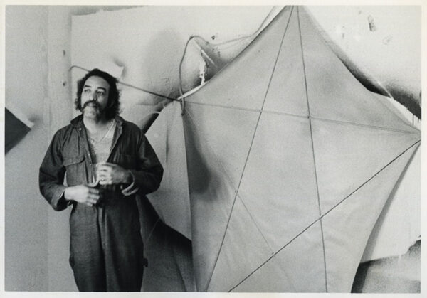 A man in a jumpsuit with a mustache stands before a large canvas painting.
