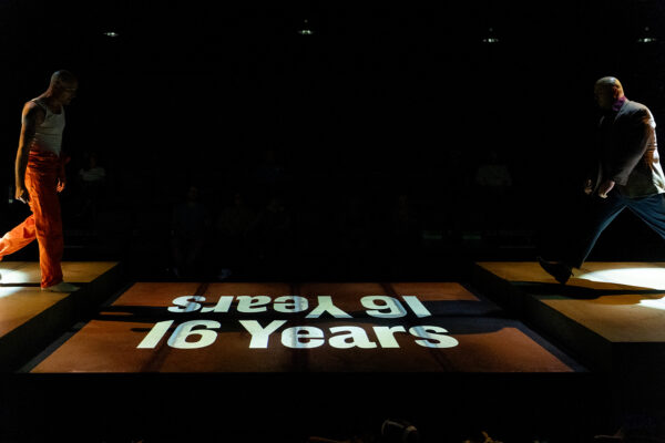 A photograph of a stage production featuring two men wearing orange, standing across the stage from each other. Text that reads, "16 Years," is projected onto the stage.