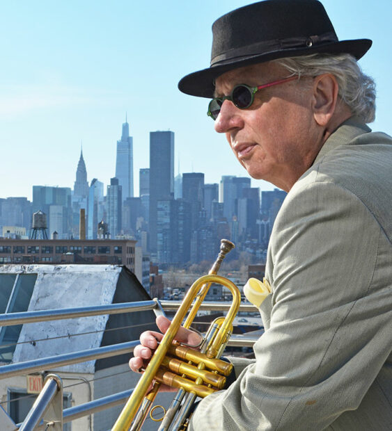 A man with a hat and sunglassess holds a trumpet in front of the New York skyline.