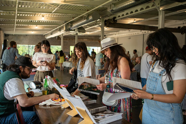Four people stand at a table of handmade photo zines in a large festival.