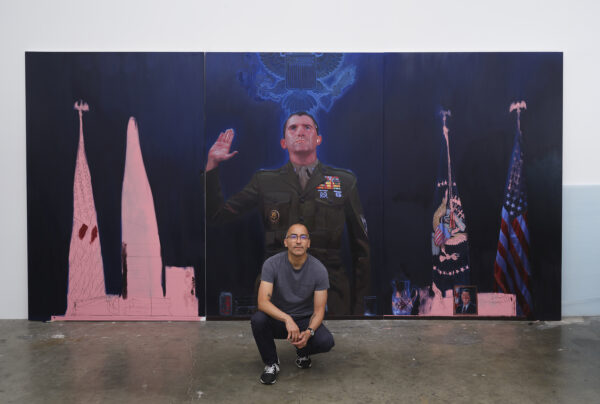 A photograph of artist Vincent Valdez in front of a large-scale painting.