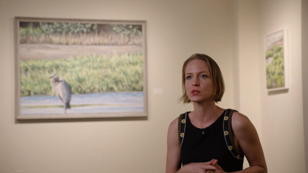 A still image of a woman standing in front of two framed artworks.