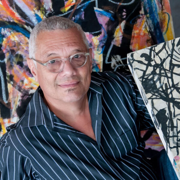 A photograph of artist Ibsen Espada surrounded by colorful abstract paintings.