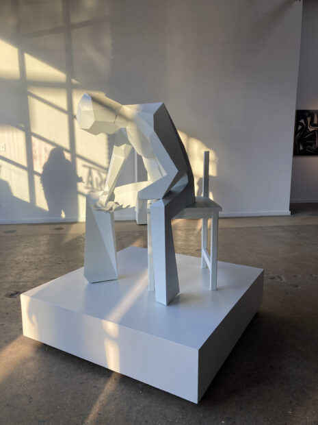A humanoid figure constructed out of polygons and painted white site on a chair and looks down toward it's face held in one hand